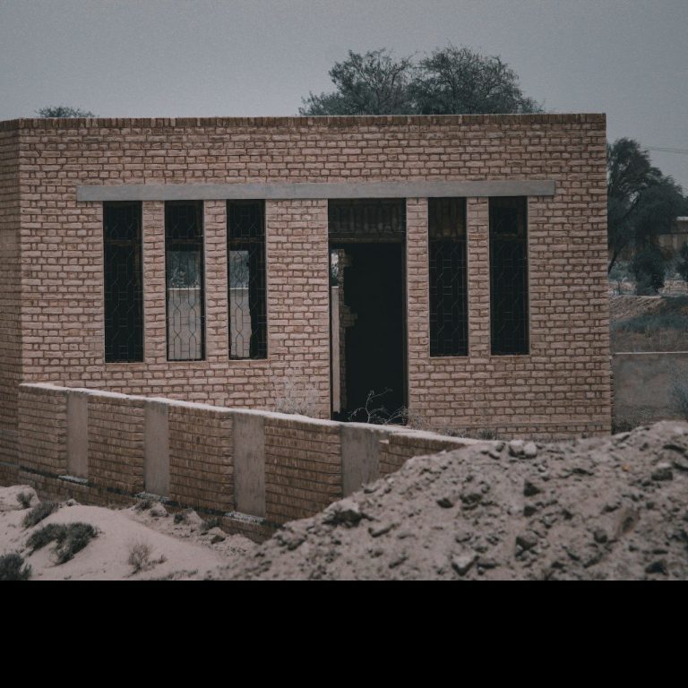 Abandoned brick building with incomplete windows, surrounded by sandy terrain.