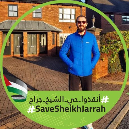 Testimonial Man in a blue jacket standing outside a brick building, holding a sign about Sheikh Jarrah.